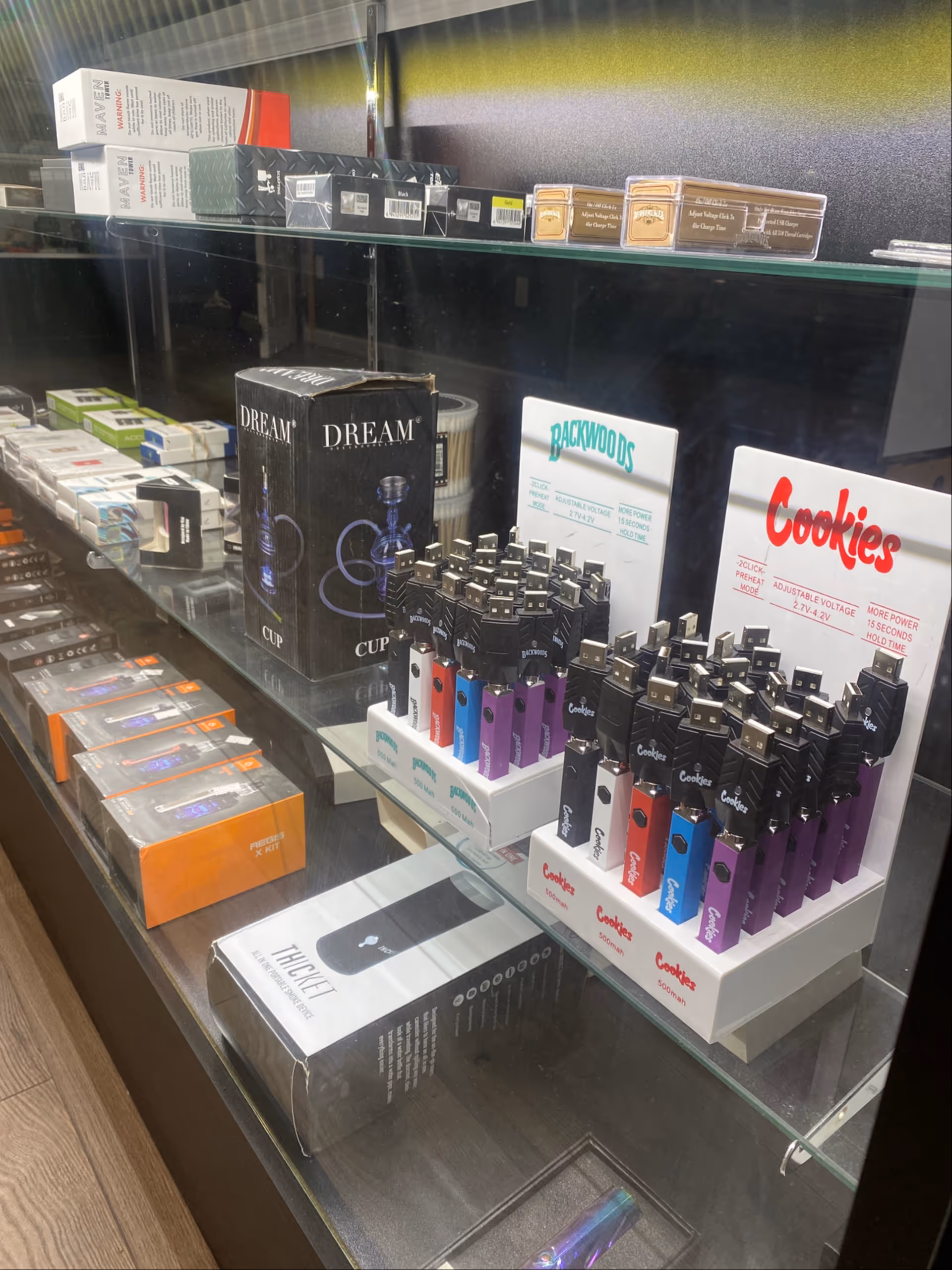 Glass display case shows assorted vape pens, boxes, and accessories in a retail shop.