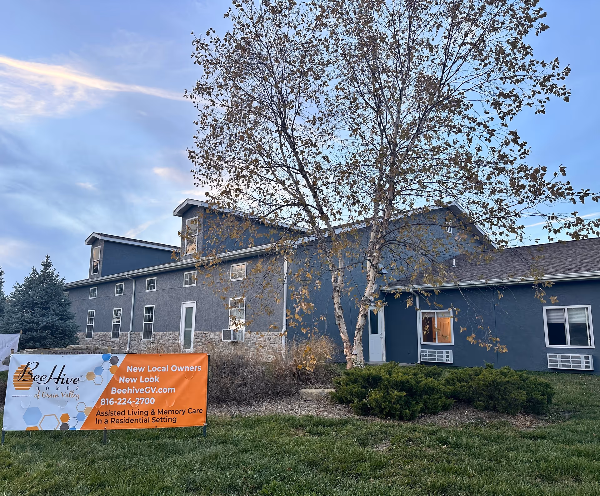 Exterior view of a two-story building with blue-gray walls and stone accents at the base. In front of the building is a tree with sparse leaves and some bushes. A large orange and white banner is displayed on the grass, advertising BeeHive Homes of Grain Valley with contact information and services for assisted living and memory care.