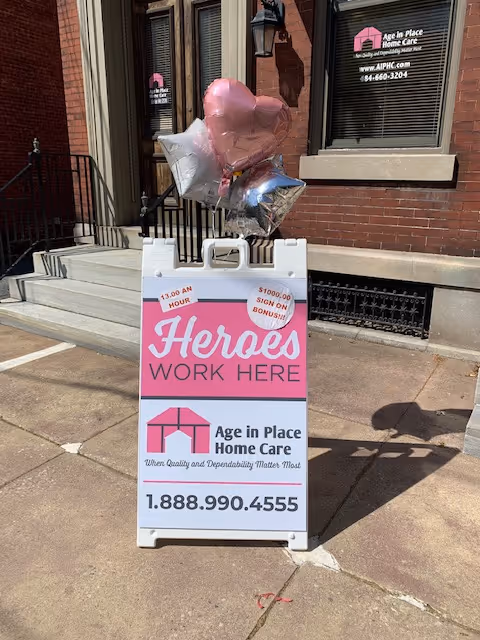Sidewalk sign for Age in Place Home Care outside a brick building with balloons.