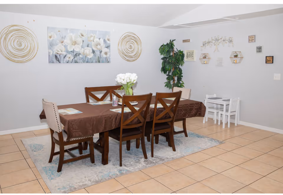 Dining room with a long table, brown tablecloth, chairs, wall art, and a plant.