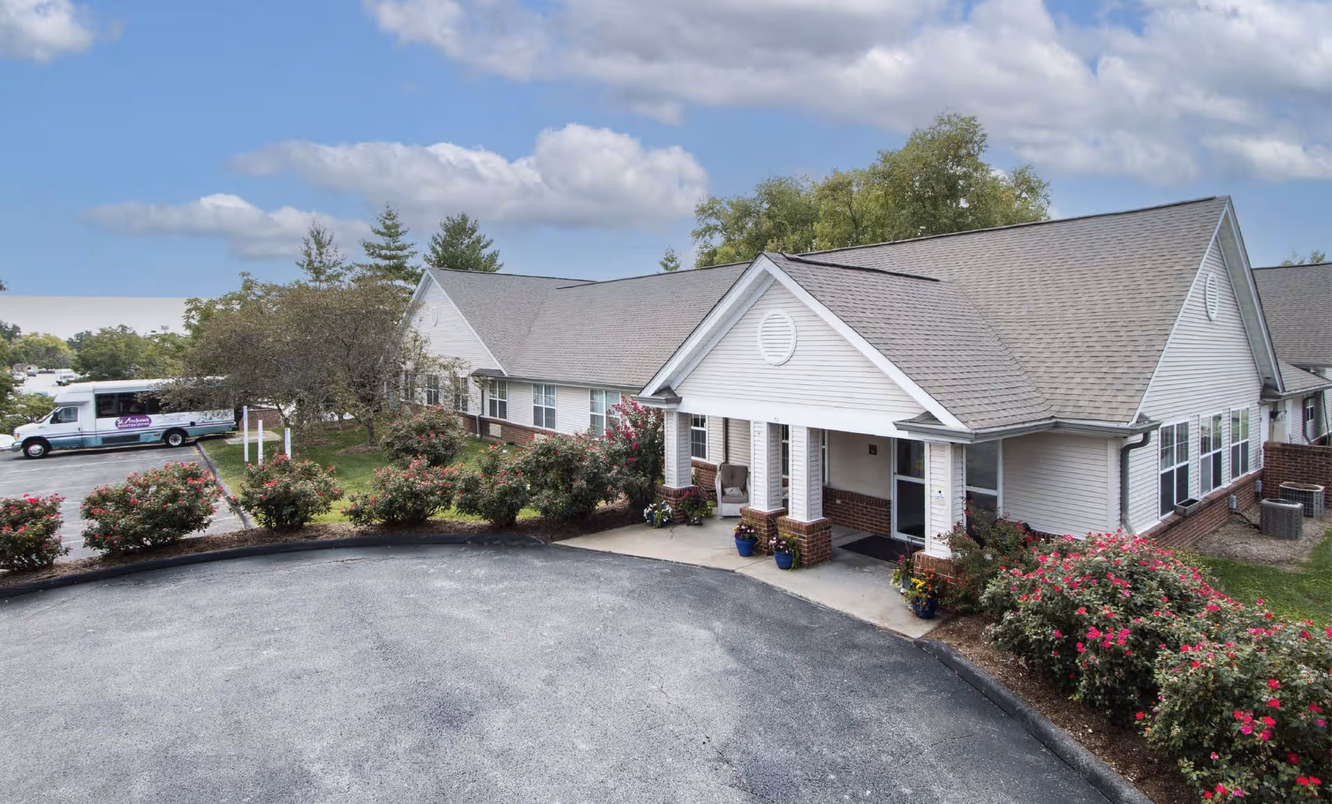 Exterior view of a single-story assisted living facility building with a covered entrance, surrounded by bushes with pink flowers and a paved driveway. A white shuttle bus is parked in the background near some trees under a partly cloudy sky.