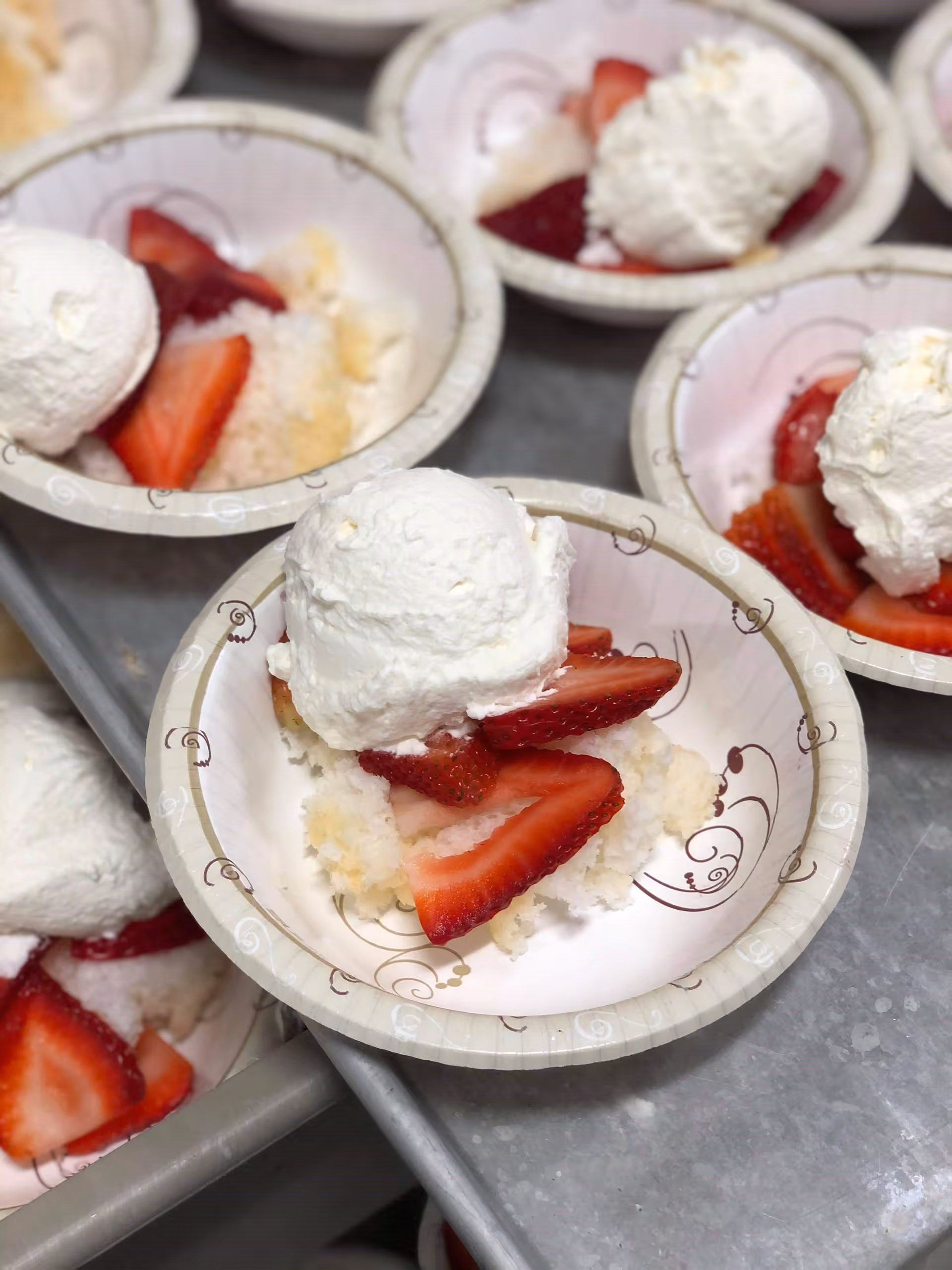 Several paper bowls each containing a dessert with sliced strawberries, a piece of cake, and a dollop of whipped cream on top, placed on a metal surface.