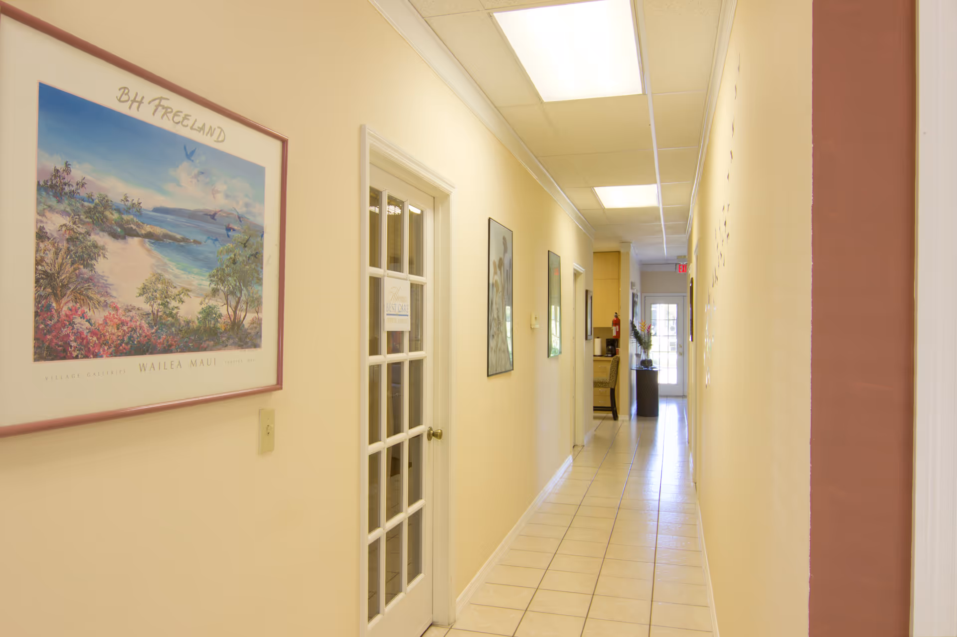 Long beige hallway with framed art, glass doors, and bright ceiling lights.