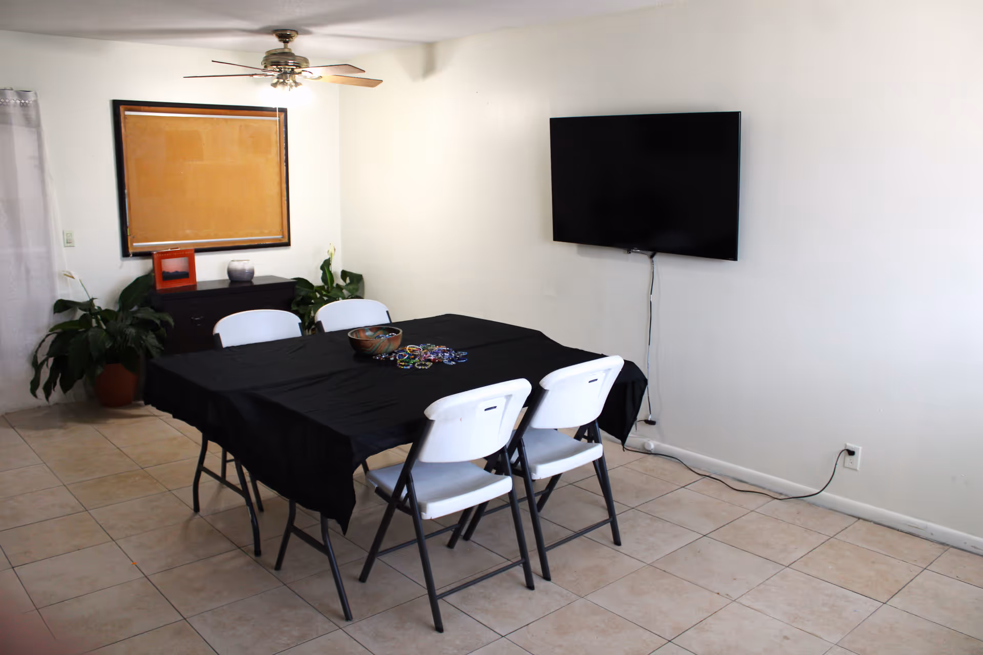 Small dining area with a black-covered table, white chairs, plants, a mounted TV, and ceiling fan.