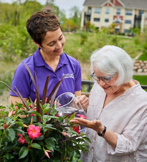 Caregiver assists elderly resident watering pink flowers in an outdoor garden near the facility.