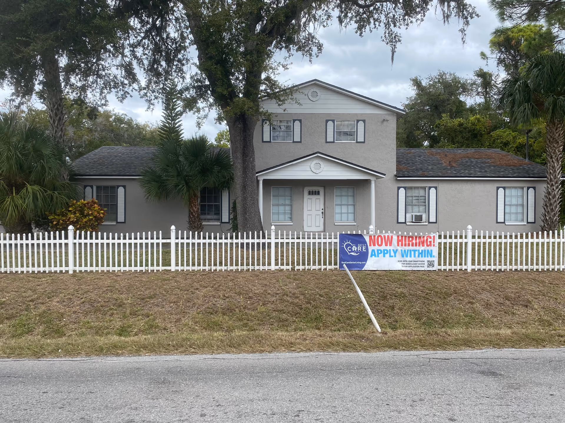 Front view of a two-story senior living building with a white picket fence and a 'Now Hiring' sign on the lawn.