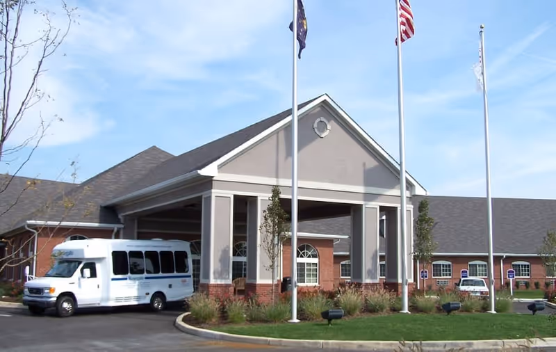 Front entrance of a senior living facility with a covered portico, three flagpoles, and a parked white shuttle van.