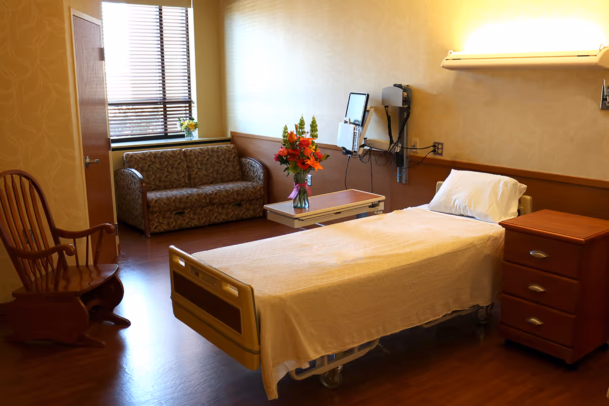 Hospital room with a single bed, couch, flowers, and medical equipment along the wall.