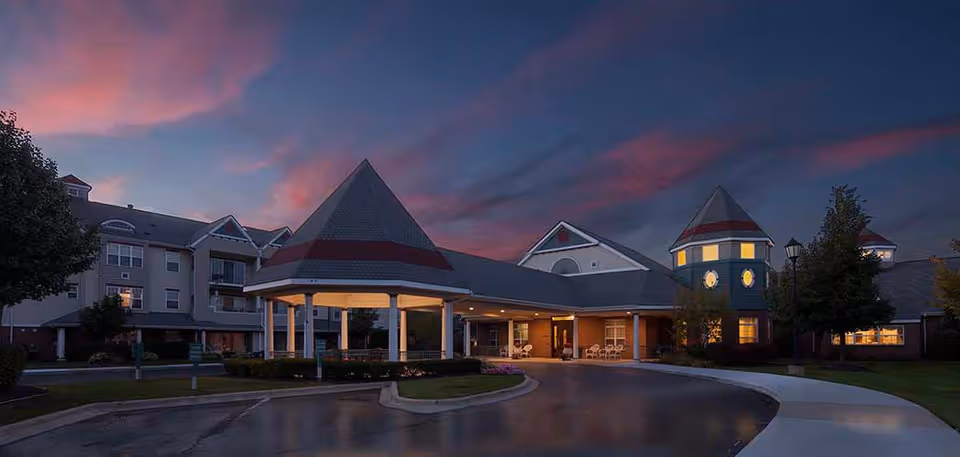 Exterior front of a senior living facility at dusk showing an illuminated covered entrance with turreted towers and a curved driveway.