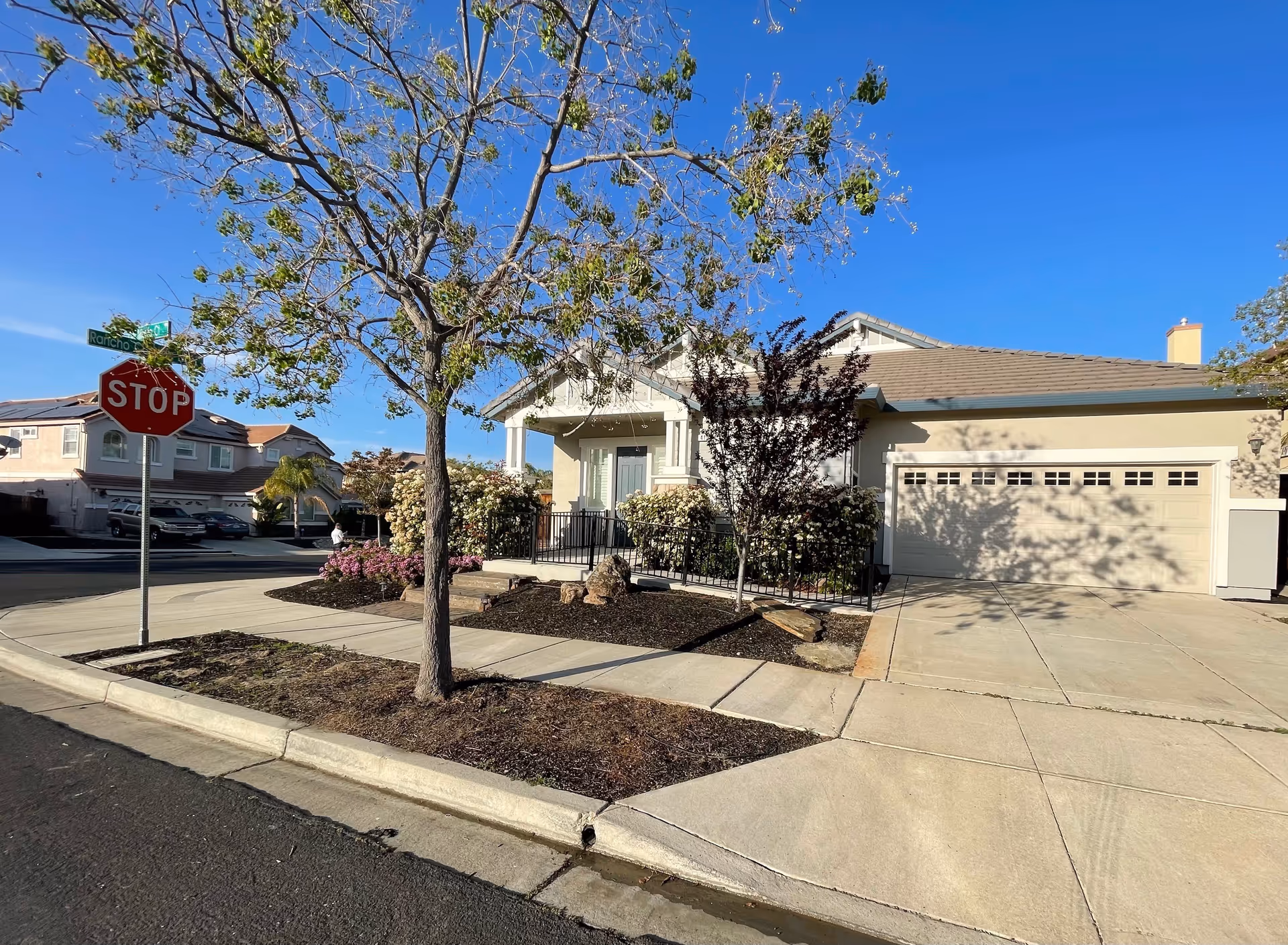 Exterior view of a single-story residential building with a beige facade and a two-car garage. The house has a small front porch with railings, surrounded by landscaped bushes and trees. A stop sign and street signs are visible on the corner of the sidewalk under a clear blue sky.