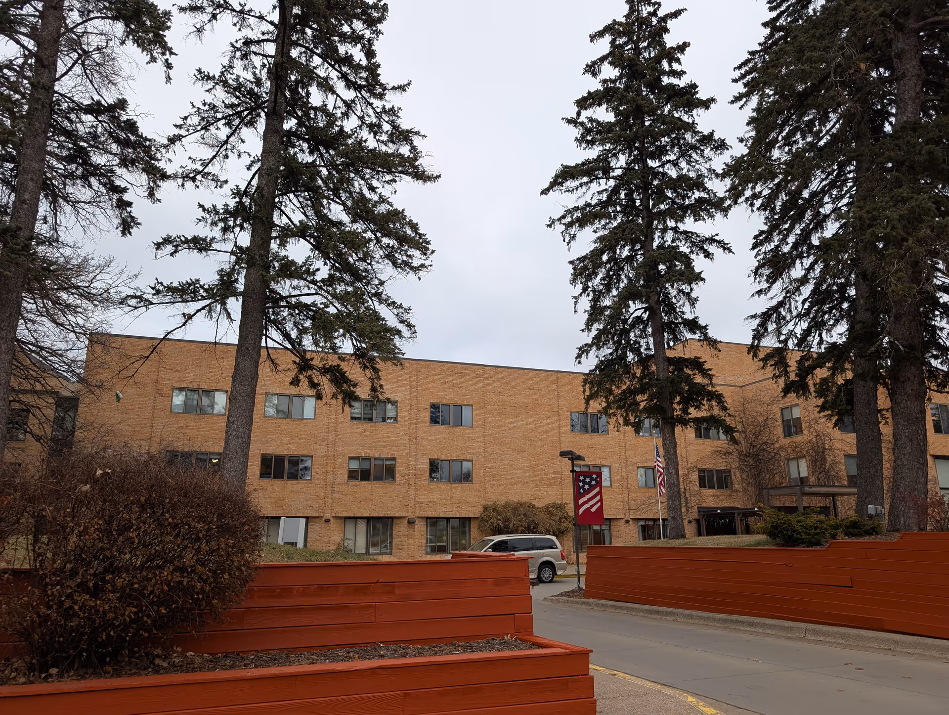 Exterior view of a three-story brick building with multiple windows, surrounded by tall evergreen trees and red wooden planters. A gray vehicle is parked near the entrance, and an American flag is displayed on a pole near the building.