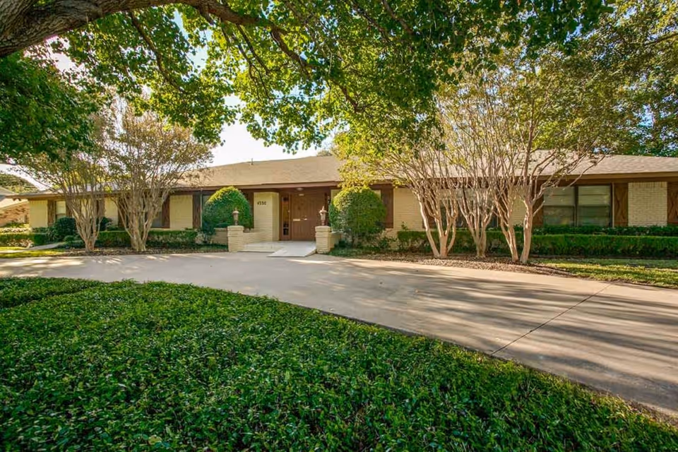 Exterior view of a single-story brick building with a sloped roof, surrounded by trees and greenery. A paved driveway leads to the entrance, which has double doors and two decorative pillars on either side. The building is partially shaded by large tree branches overhead.