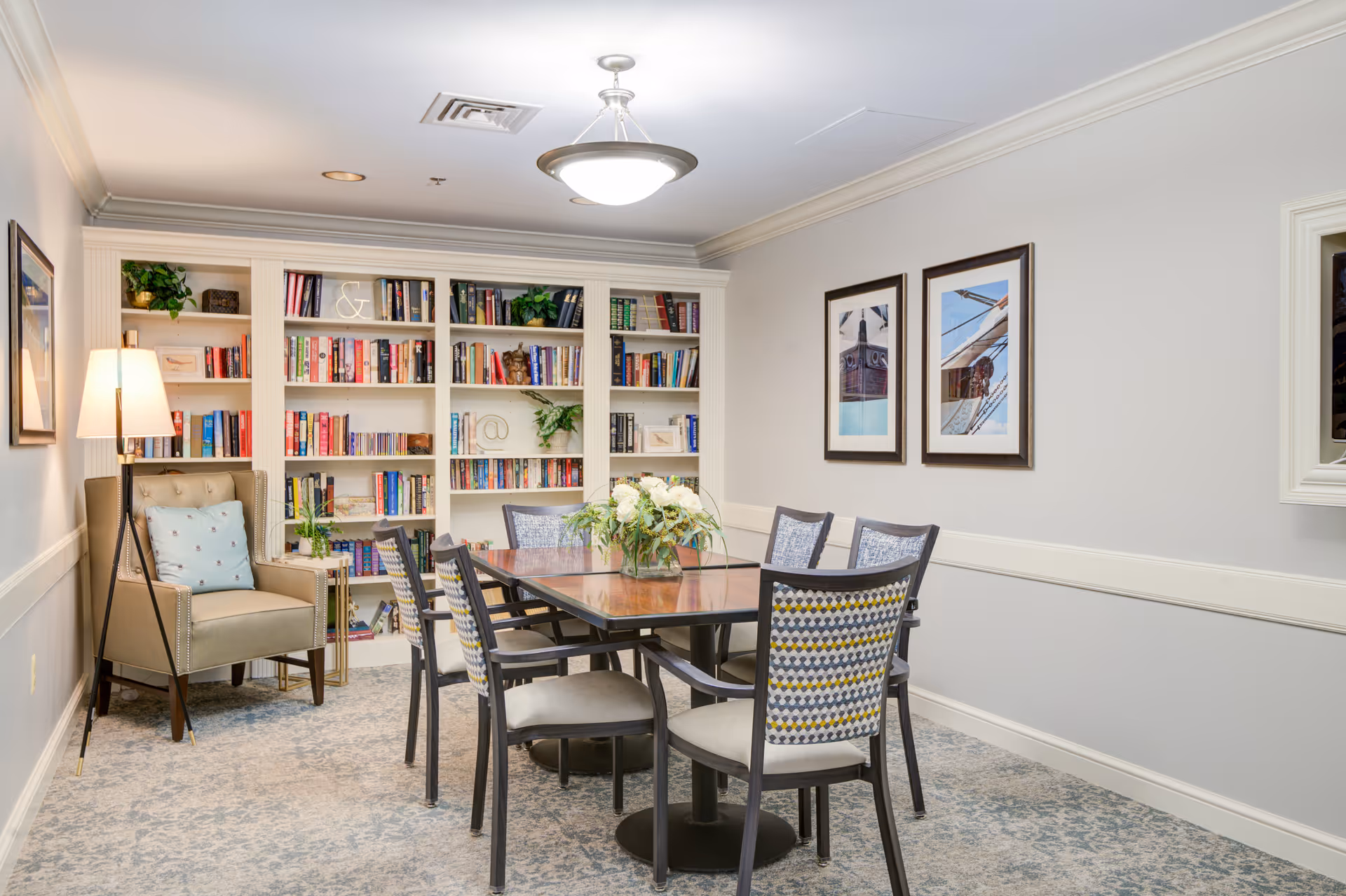 A cozy communal room with a central table and chairs, bookshelves along the back wall, an armchair and framed artwork.