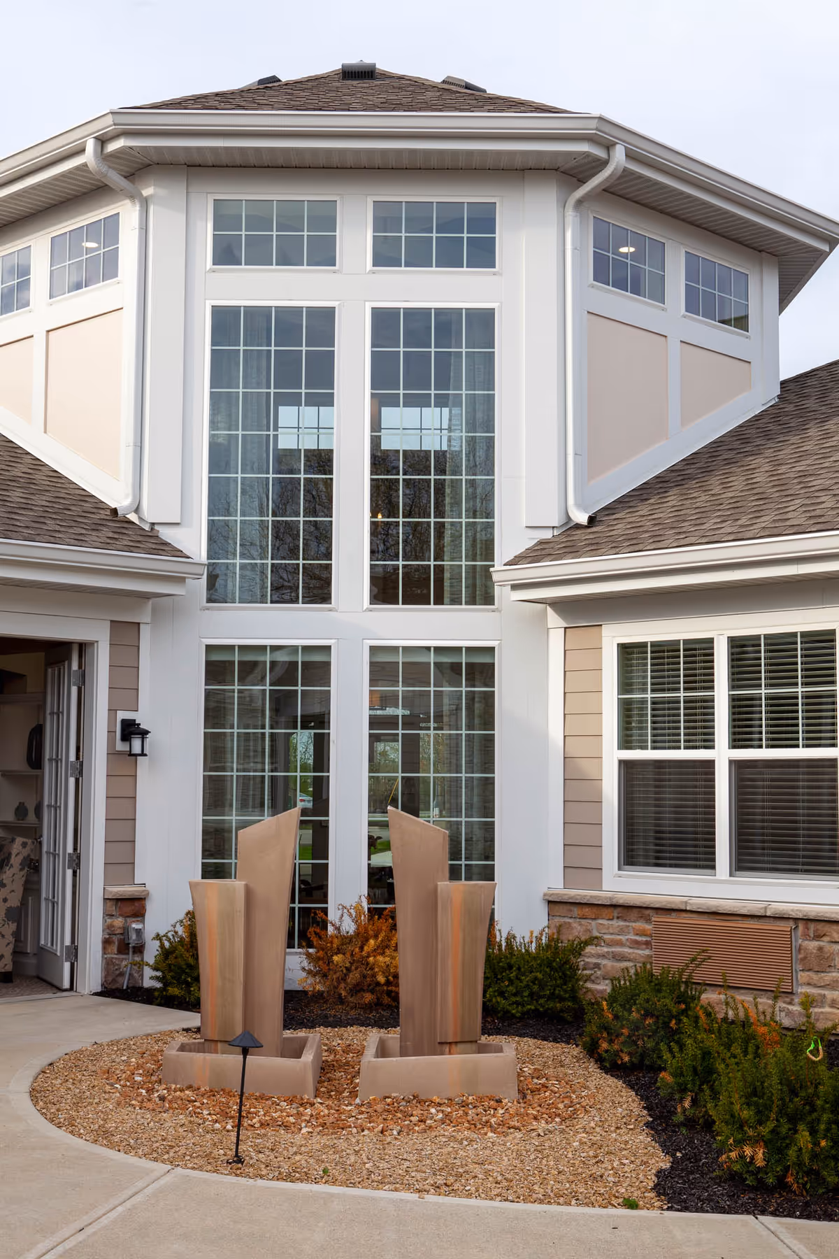Exterior front of a senior living facility featuring tall multi-pane windows, a central glass entrance, and two vertical metal sculptures in a gravel planting bed.