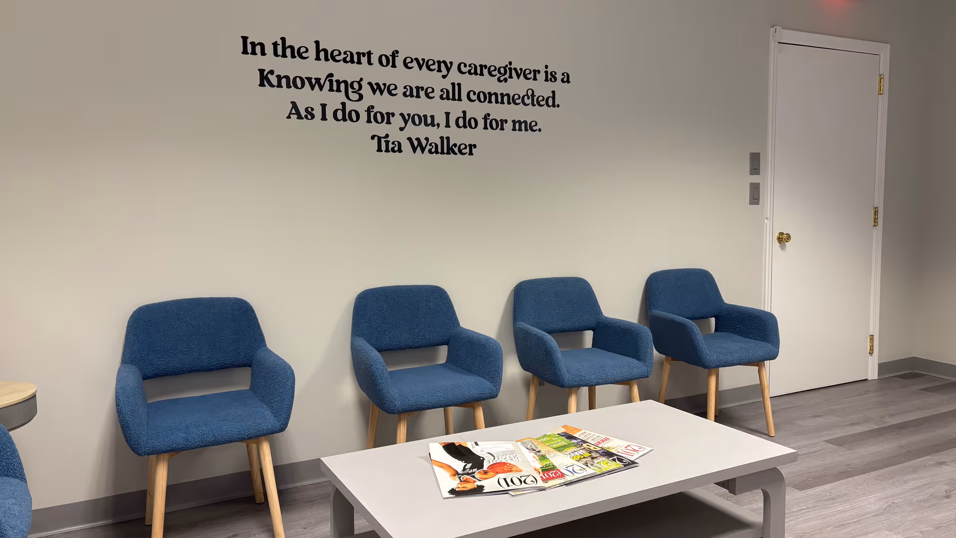 Row of blue chairs along a light wall, coffee table with magazines, and a doorway.