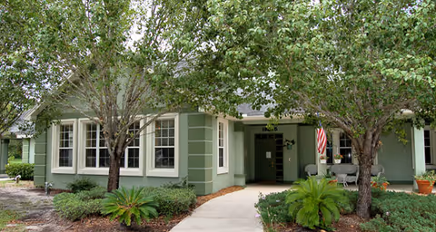 Exterior view of a single-story green building with white trim, surrounded by trees and shrubs. A concrete pathway leads to the entrance, which has an American flag displayed near the door.