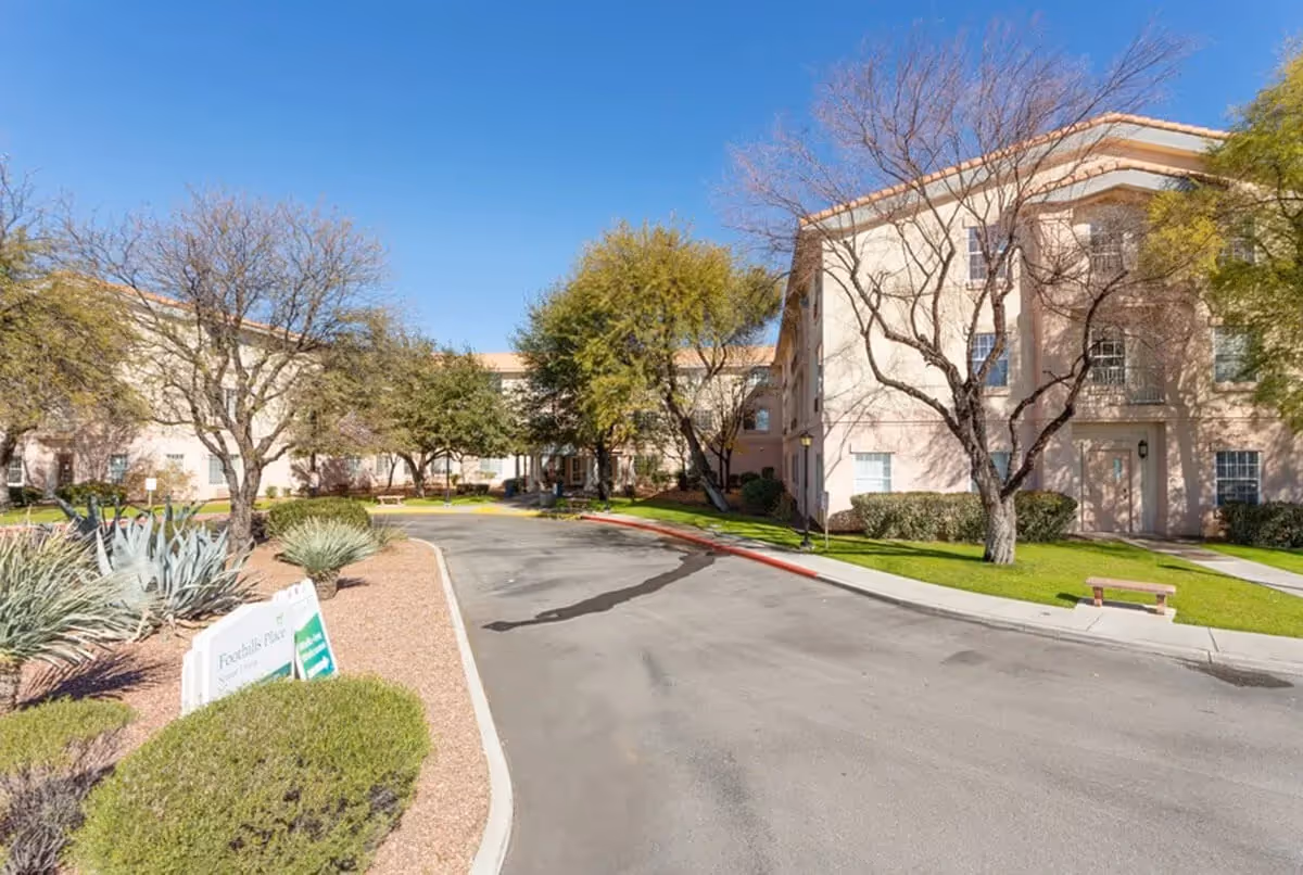 Driveway and landscaped front entrance of a three-story senior living building with trees, shrubs, a bench, and a small sign.