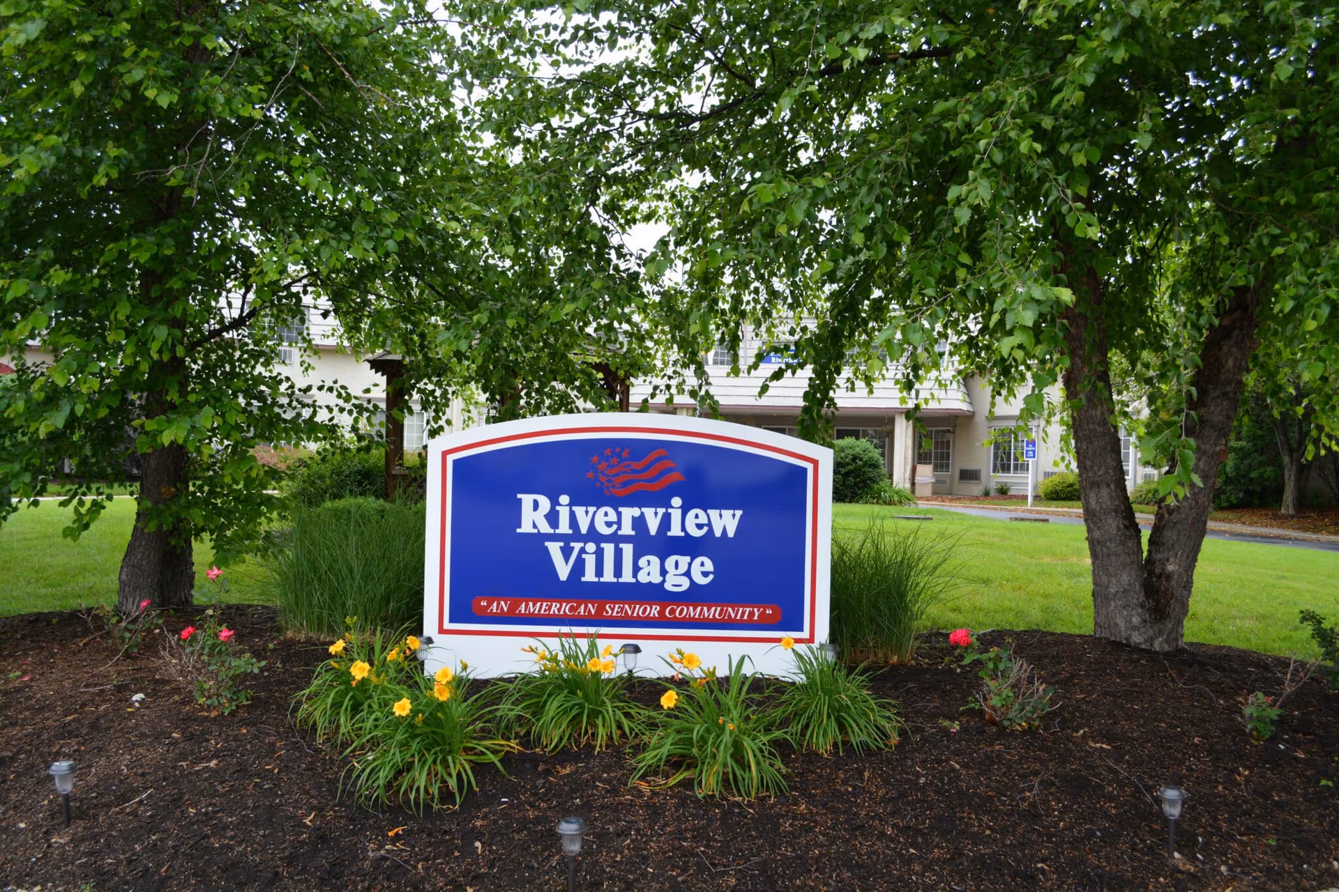 A blue and white 'Riverview Village' community sign surrounded by flowers, mulch and trees with a building visible in the background.