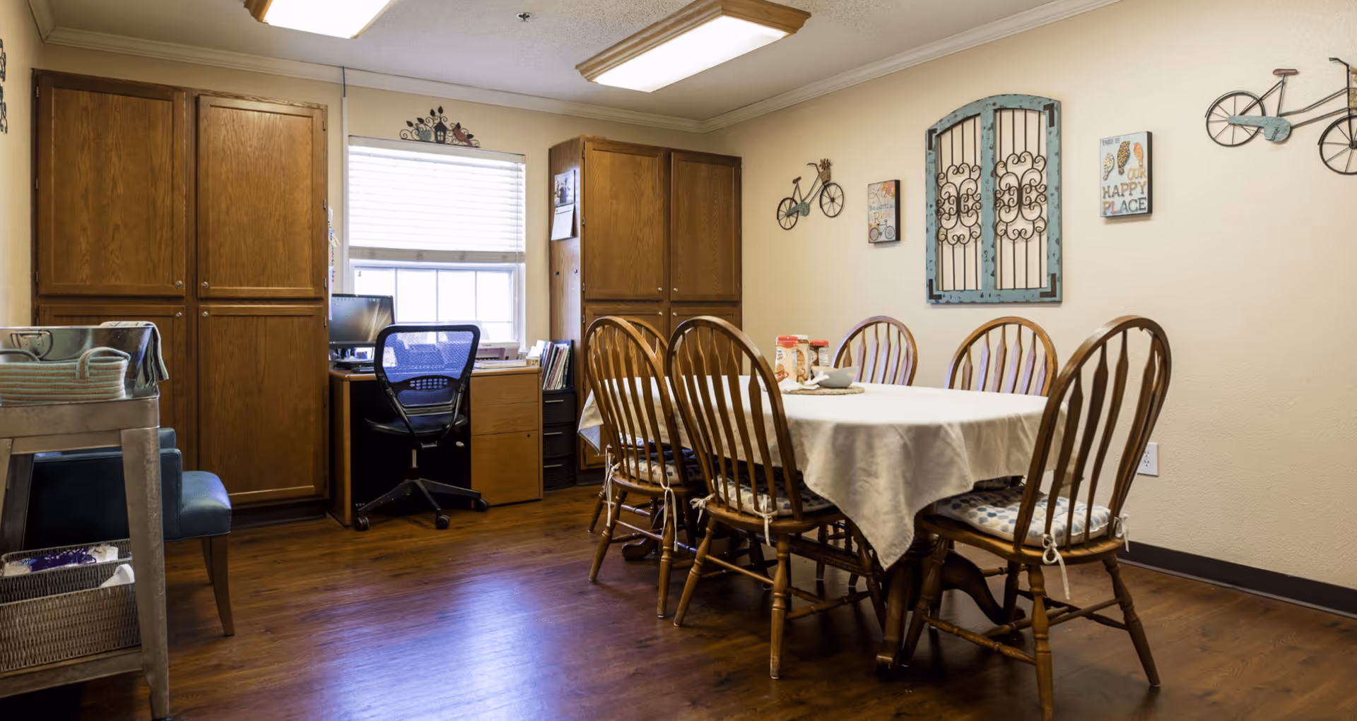 Dining room with a rectangular table covered by a white tablecloth surrounded by wooden chairs, wooden cabinets and a desk by the window, with decorative wall art.