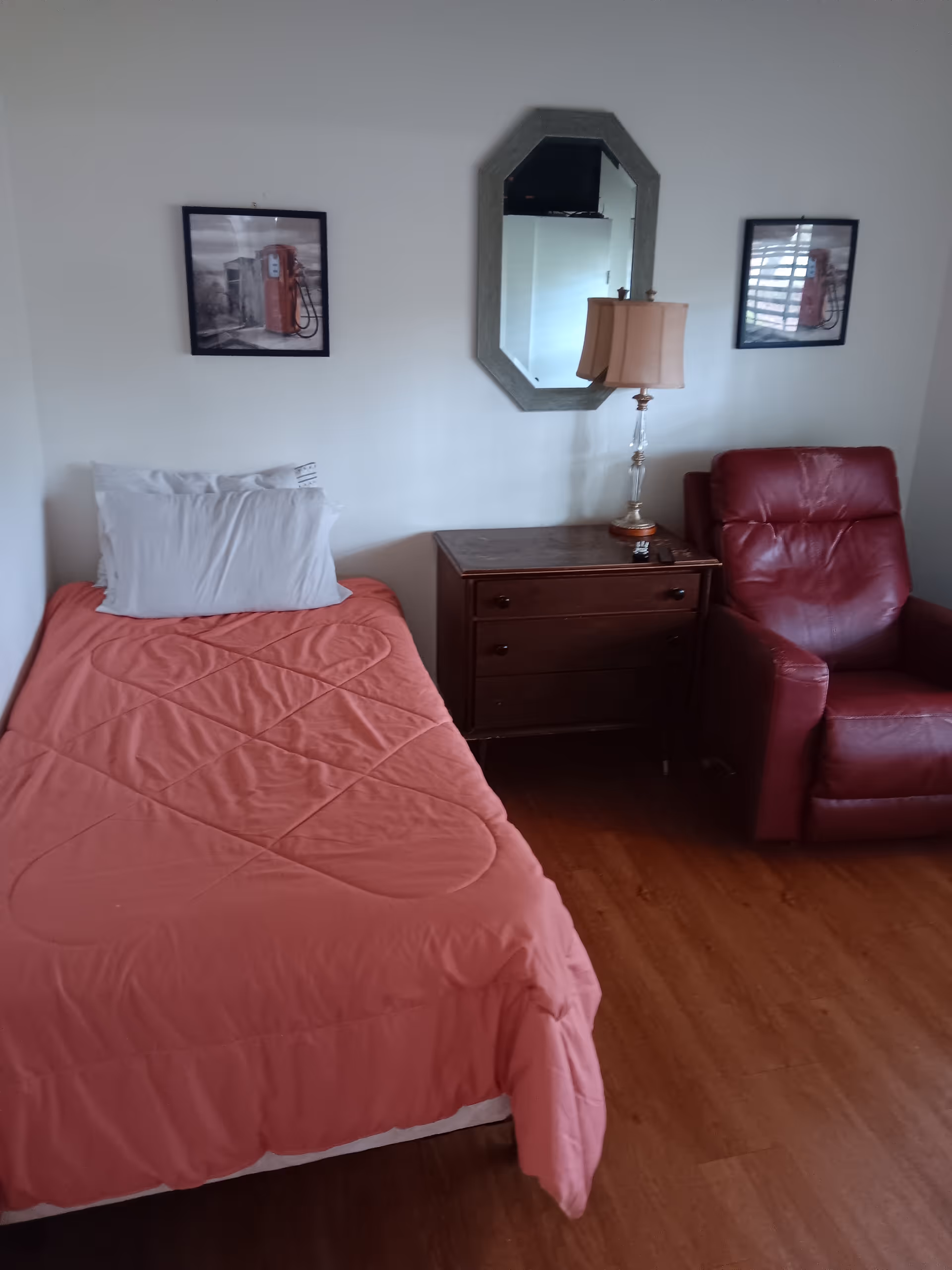 A small bedroom featuring a pink bedspread, nightstand, lamp, mirror, and red chair.