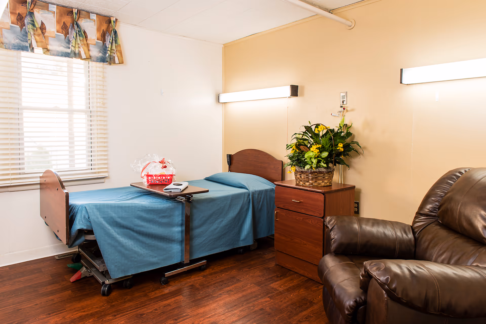 A small room with a single hospital-style bed covered with a blue blanket, a wooden nightstand with a basket of yellow flowers, a brown leather recliner chair, and a window with blinds and a patterned valance.