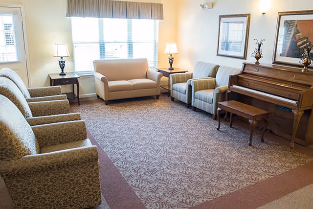 Bright common living room with upholstered chairs, a loveseat, side tables, lamps and an upright piano near a window.