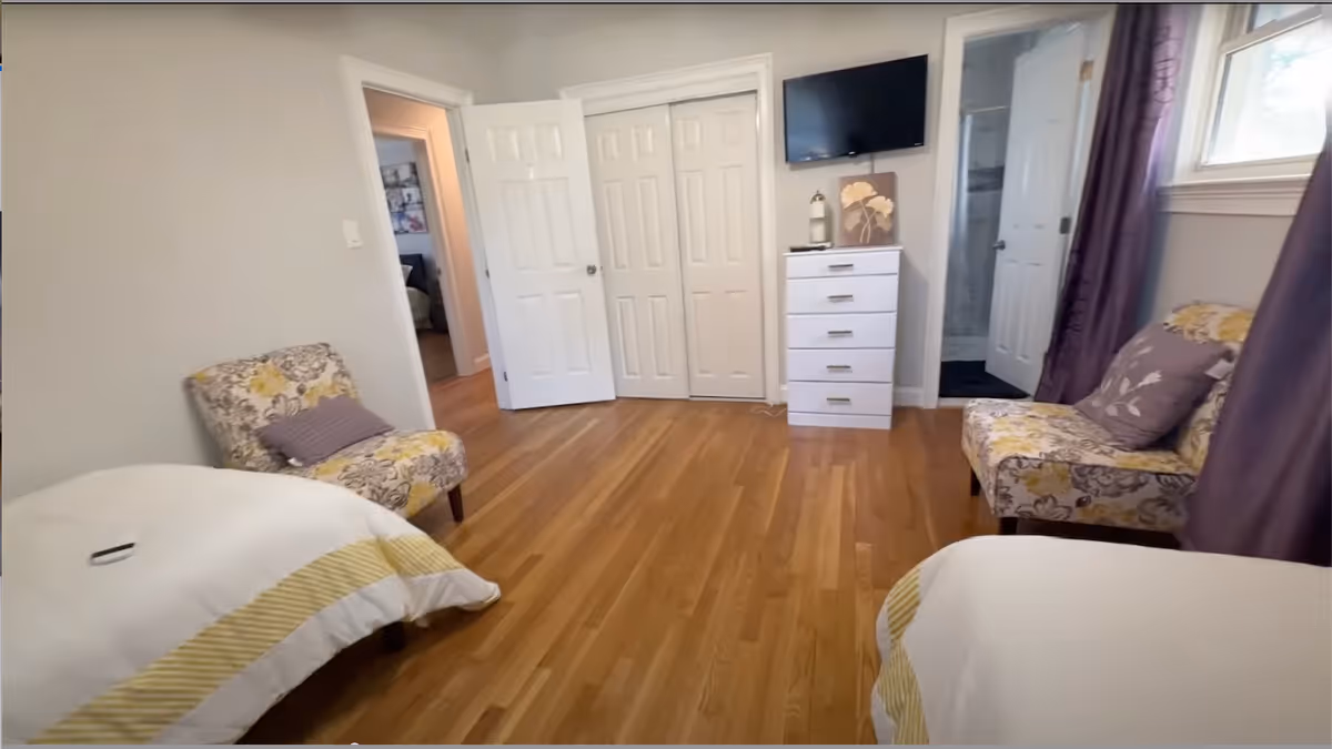 Two beds and floral chairs fill a bright senior living bedroom with a dresser and TV.