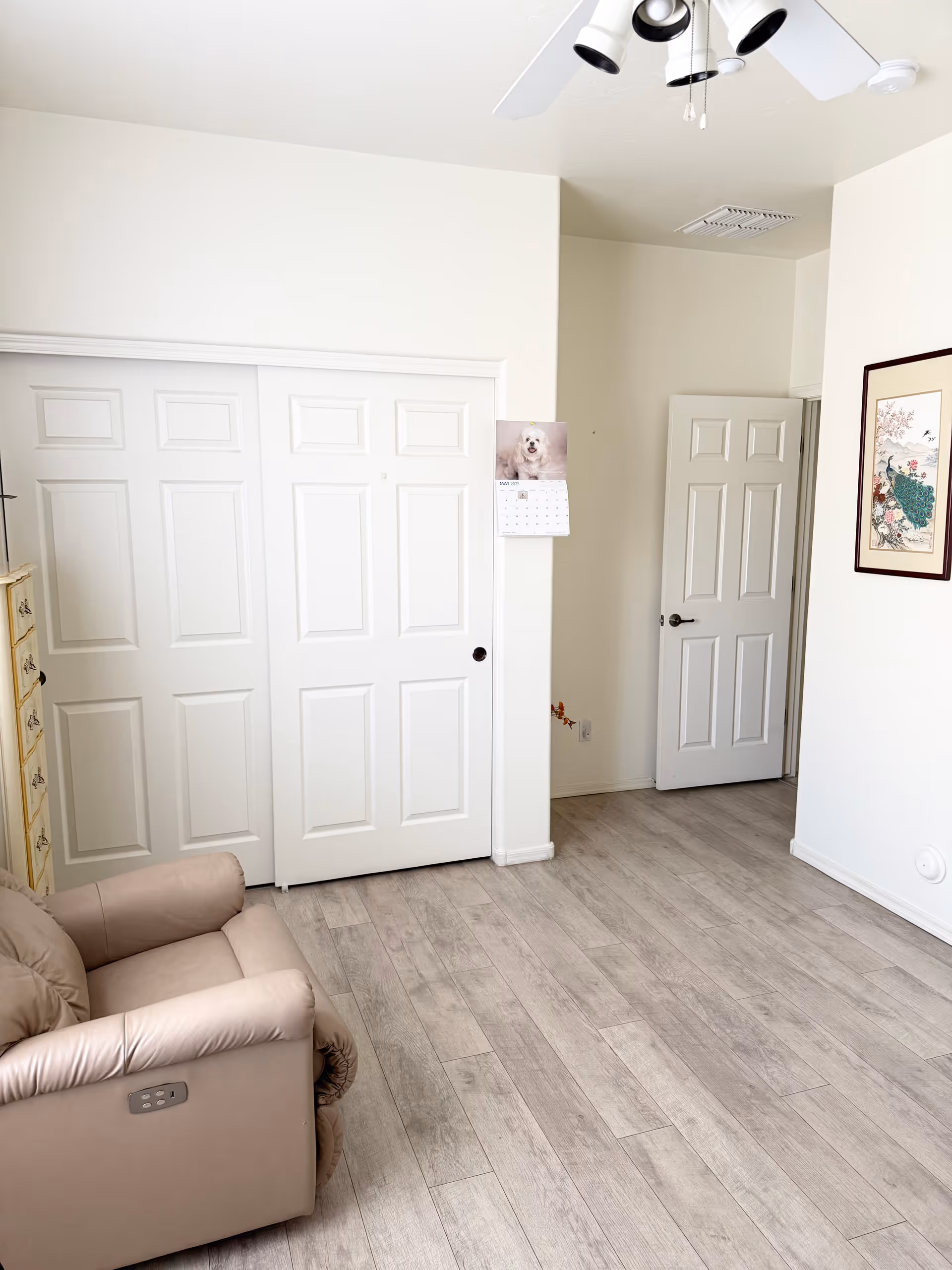 Bright beige interior featuring a recliner, sliding closet doors, calendar, and framed art.