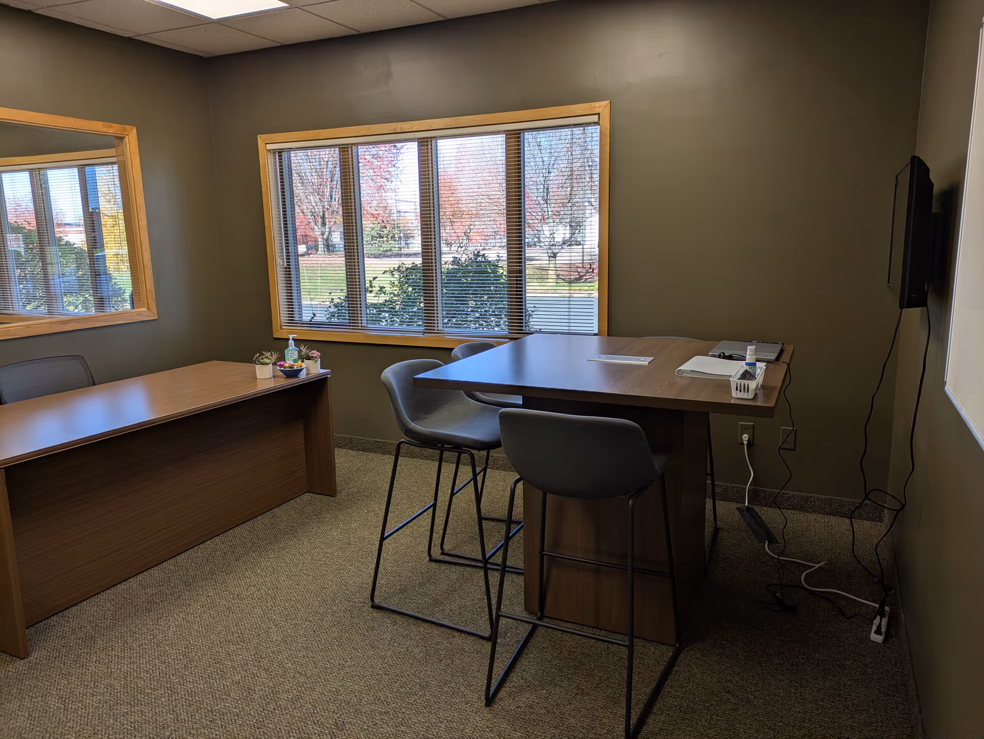 Interior meeting room with two wooden desks, chairs, windows, and a wall-mounted TV.
