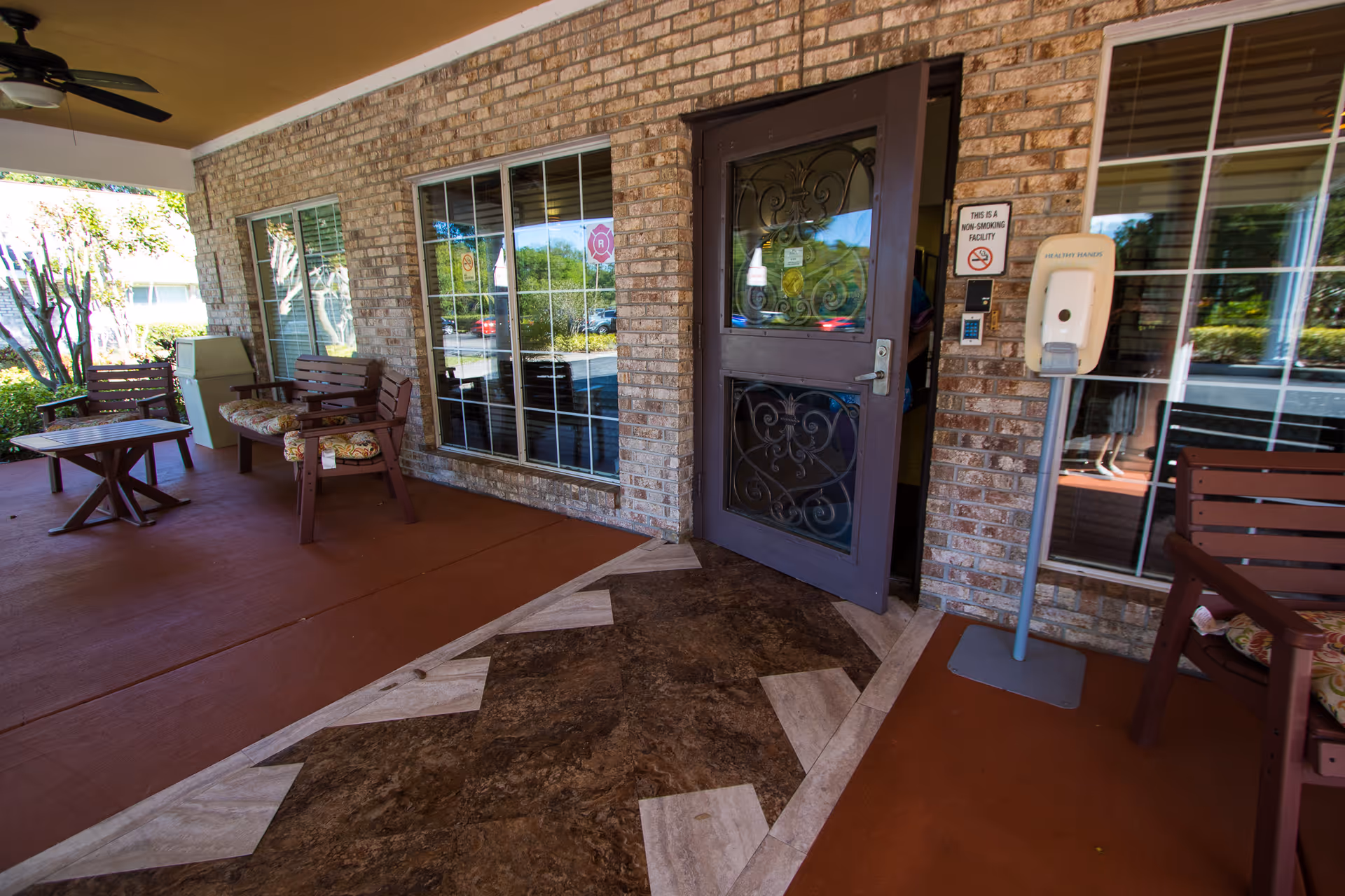 Covered brick front porch with an open decorative entry door, benches and a hand-sanitizer dispenser.