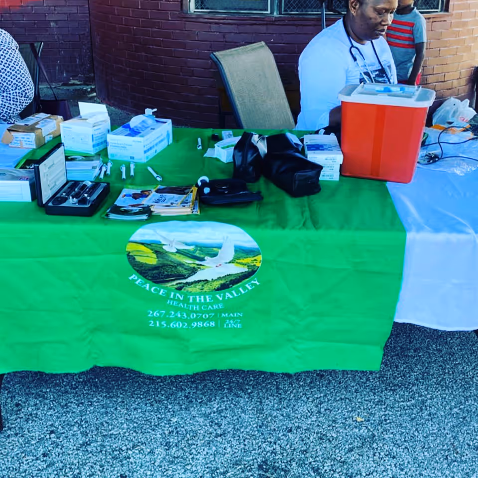 Outdoor health fair booth with a green tablecloth logo, medical supplies, cooler, and staff.