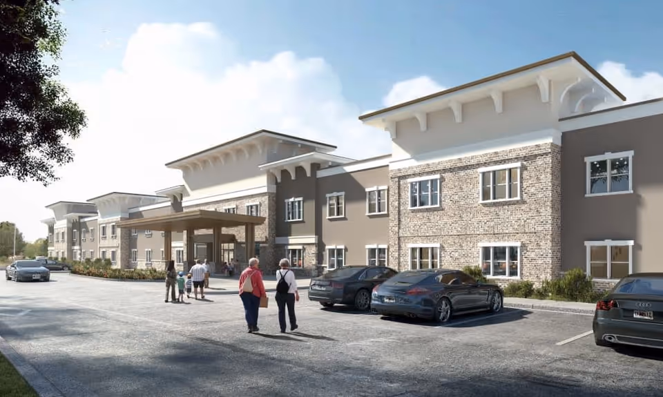 Exterior view of a modern two-story senior living facility with a covered entrance. Several people, including seniors and children, are walking towards the entrance. Multiple cars are parked in front of the building under a clear sky.