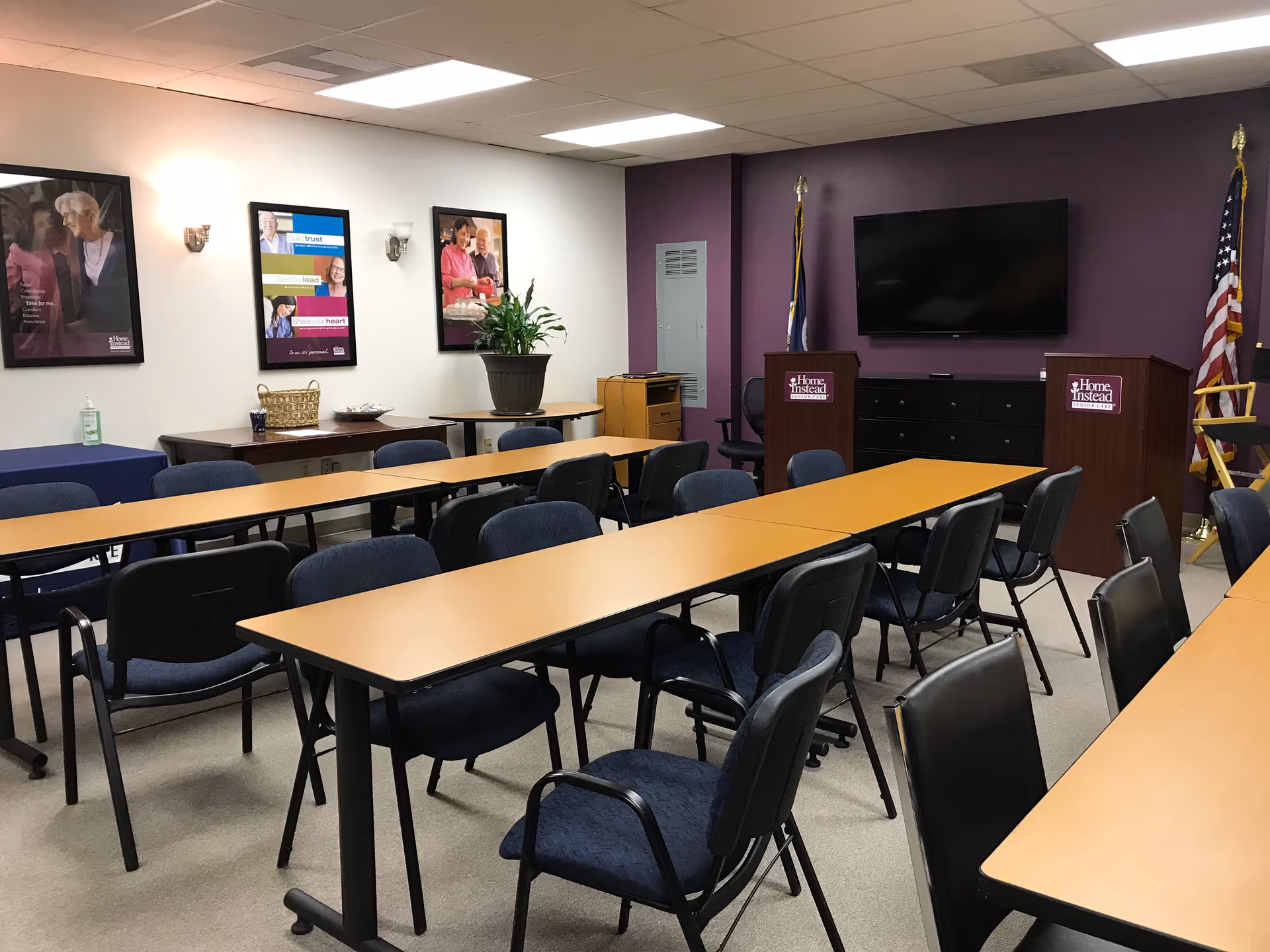 Conference-style room with long tables, many chairs, podiums, TV, purple accent wall, and posters.