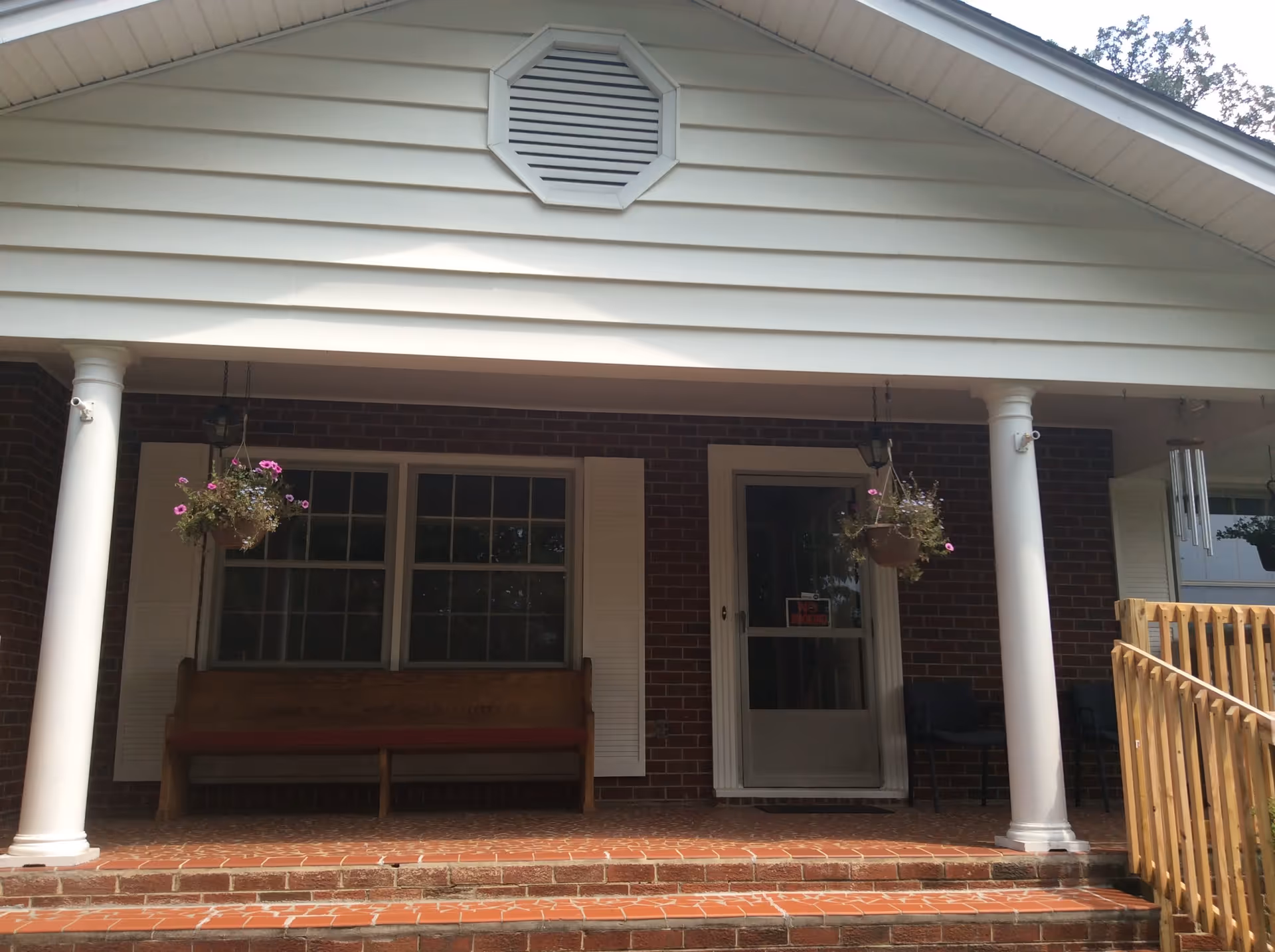 Front porch of a brick building with white columns, hanging flower baskets, a bench and a door reached by brick steps.