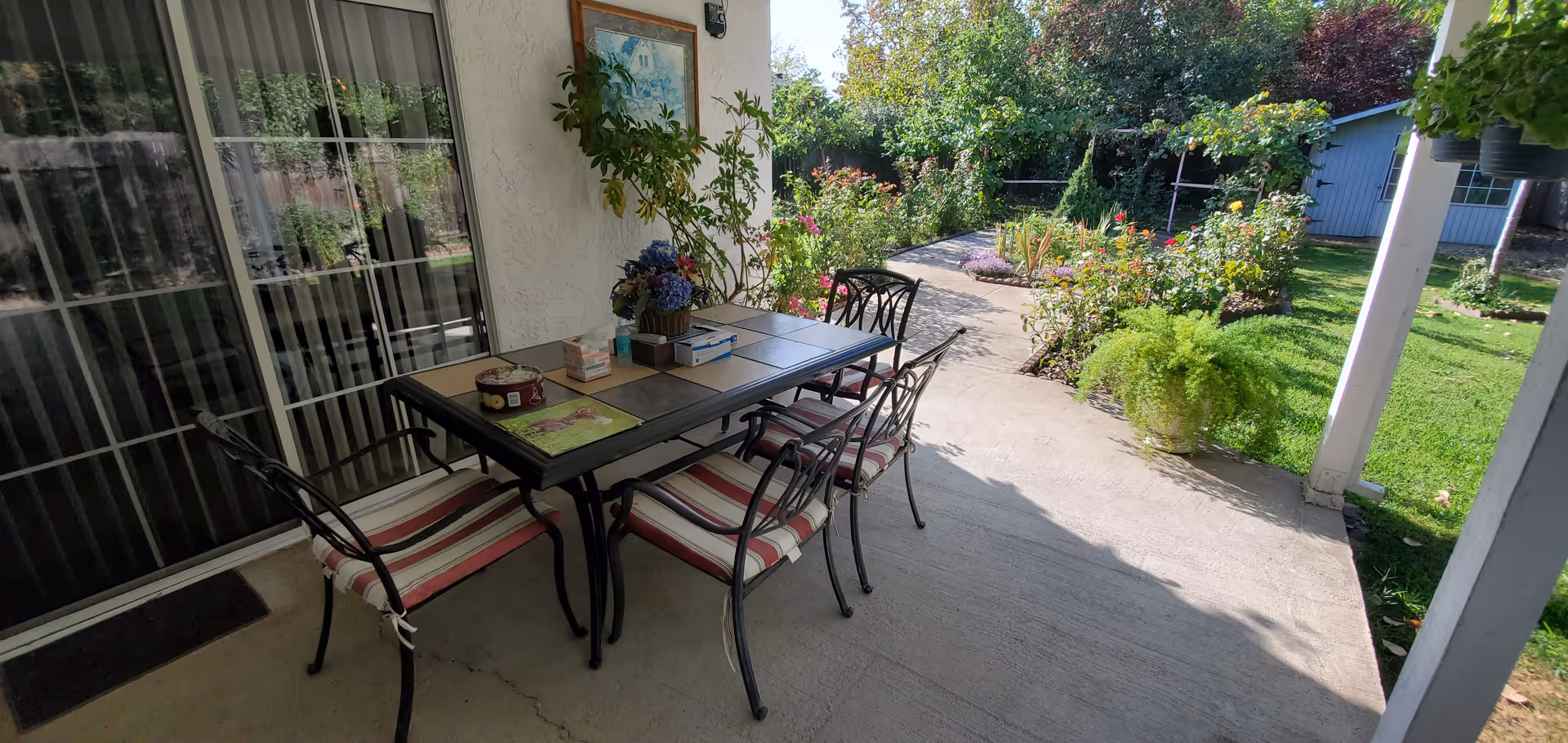 Outdoor patio table with striped cushioned chairs on a concrete porch, beside a lush garden.
