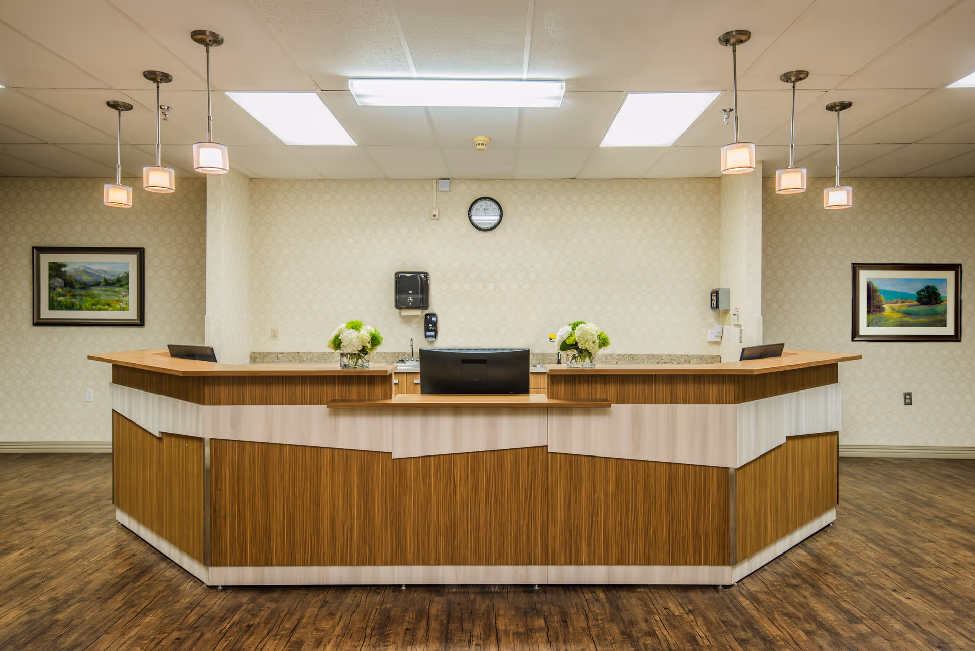 A large wood-paneled reception desk in a bright lobby with hanging pendant lights, floral arrangements, and framed artwork on the walls.