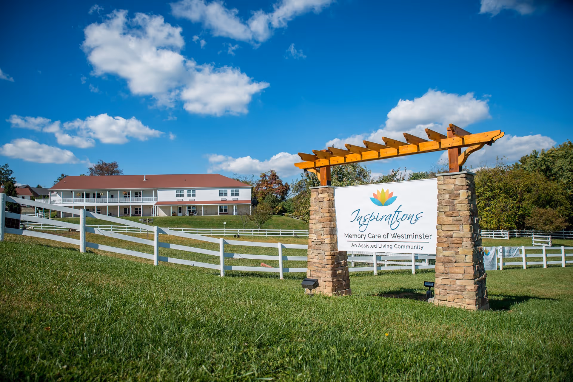 A front entrance sign reading 'Inspirations Memory Care of Westminster' on stone pillars in front of a white, two-story assisted living building and fenced lawn under a blue sky.
