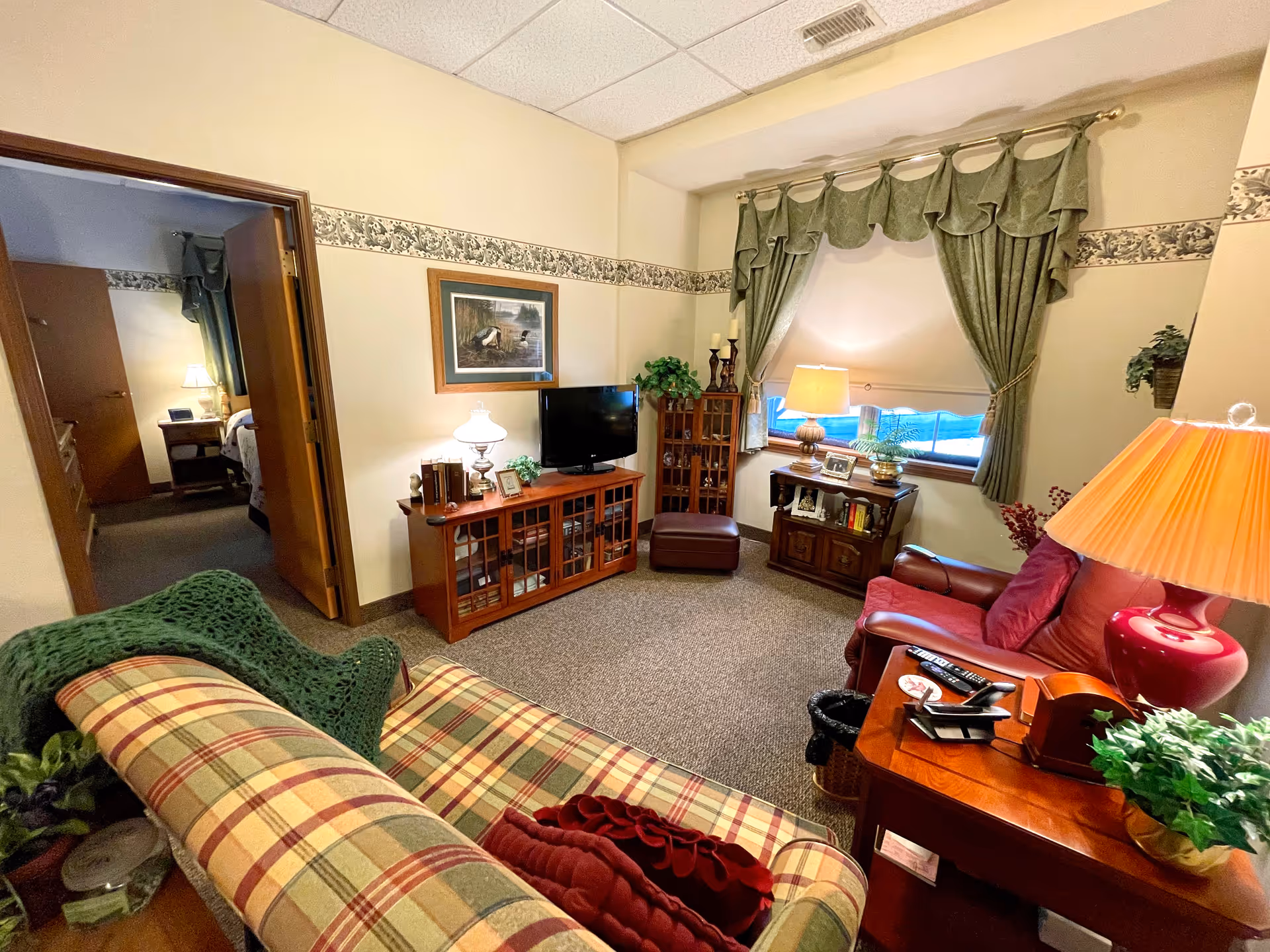A cozy living room with a plaid sofa, a red leather armchair, wooden furniture including a TV stand with a flat-screen TV, a small cabinet, and a side table with a red lamp. The room has beige walls with a decorative border, a window with green curtains, and a doorway leading to a bedroom.
