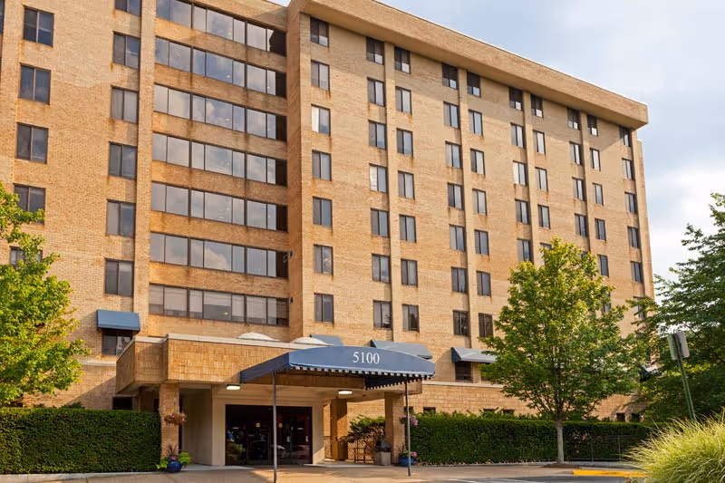 Front exterior of a multi-story brick senior living building with an entrance canopy marked "5100" and surrounding trees and landscaping.