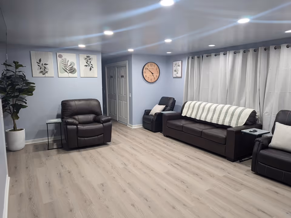 Light blue living room with leather sofas, recliners, clock, plants, wall art, and white curtains.