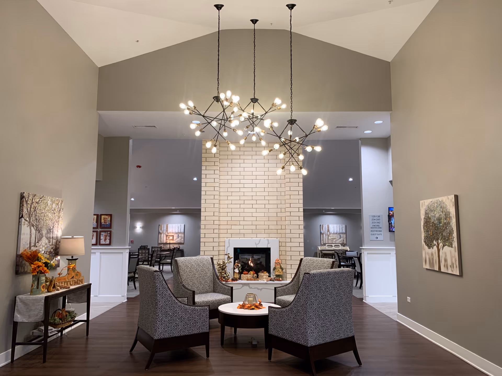 A spacious modern senior living lounge with a brick fireplace, four armchairs, and dramatic chandeliers.