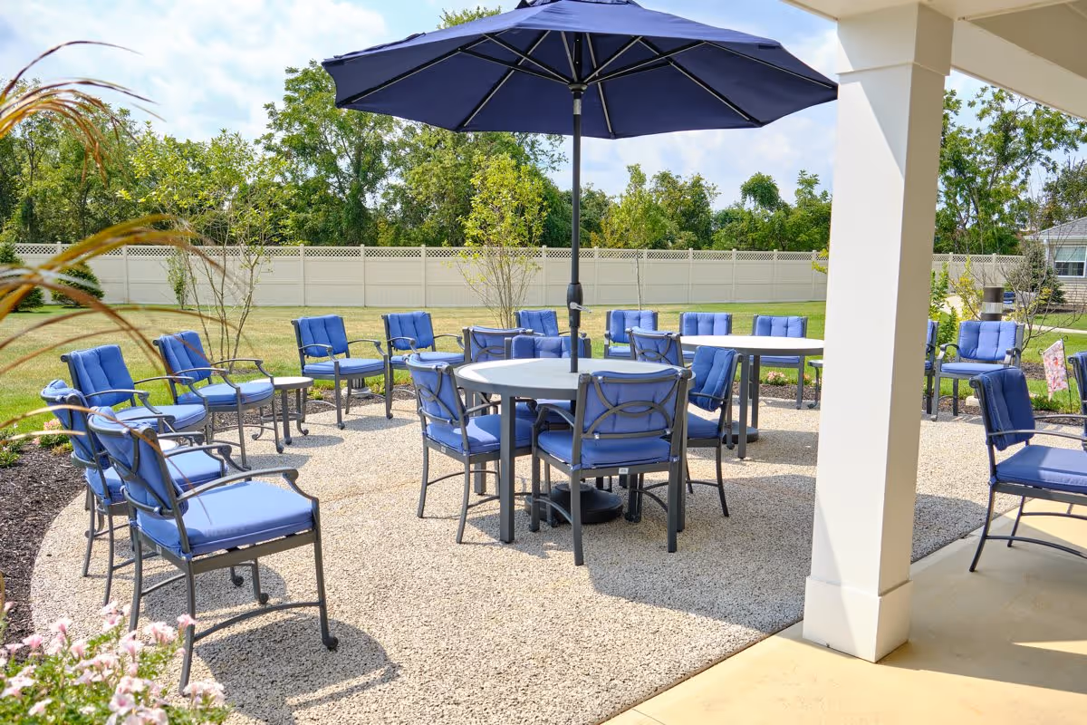 Outdoor patio area with multiple blue cushioned chairs arranged around circular tables, one table shaded by a large blue umbrella. The patio is surrounded by a grassy yard with trees and a white fence in the background.