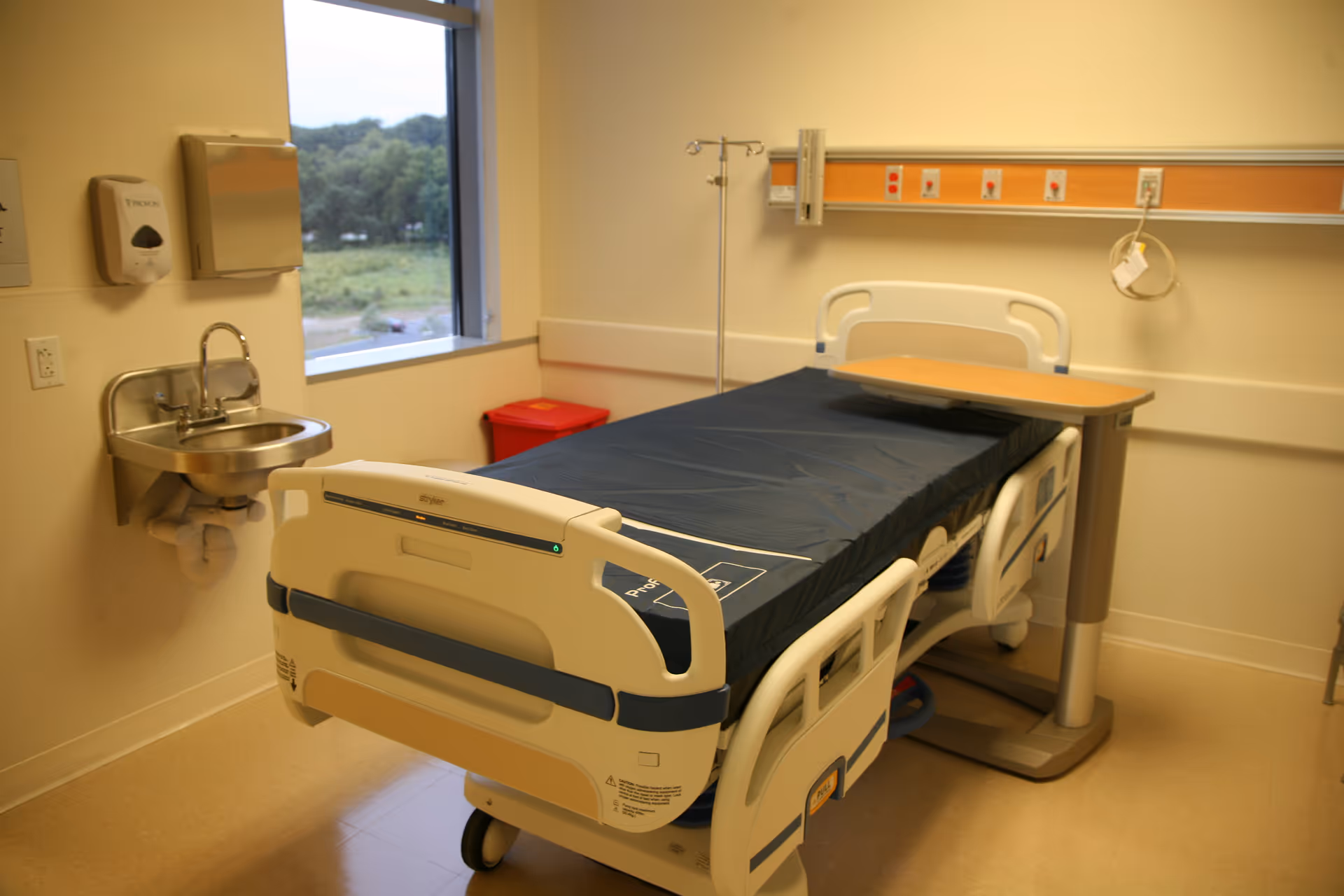 Hospital-style patient bed with side rails, table, sink, and medical equipment in a clinical room.