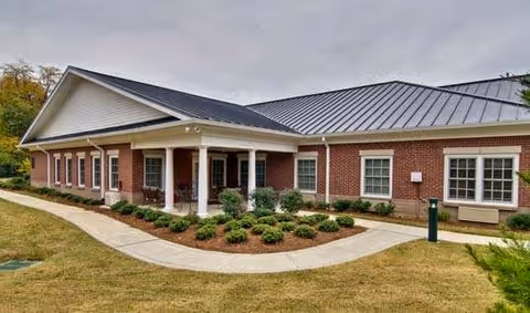 Brick senior living facility with a curved concrete walkway and landscaped entrance under a gray sky.