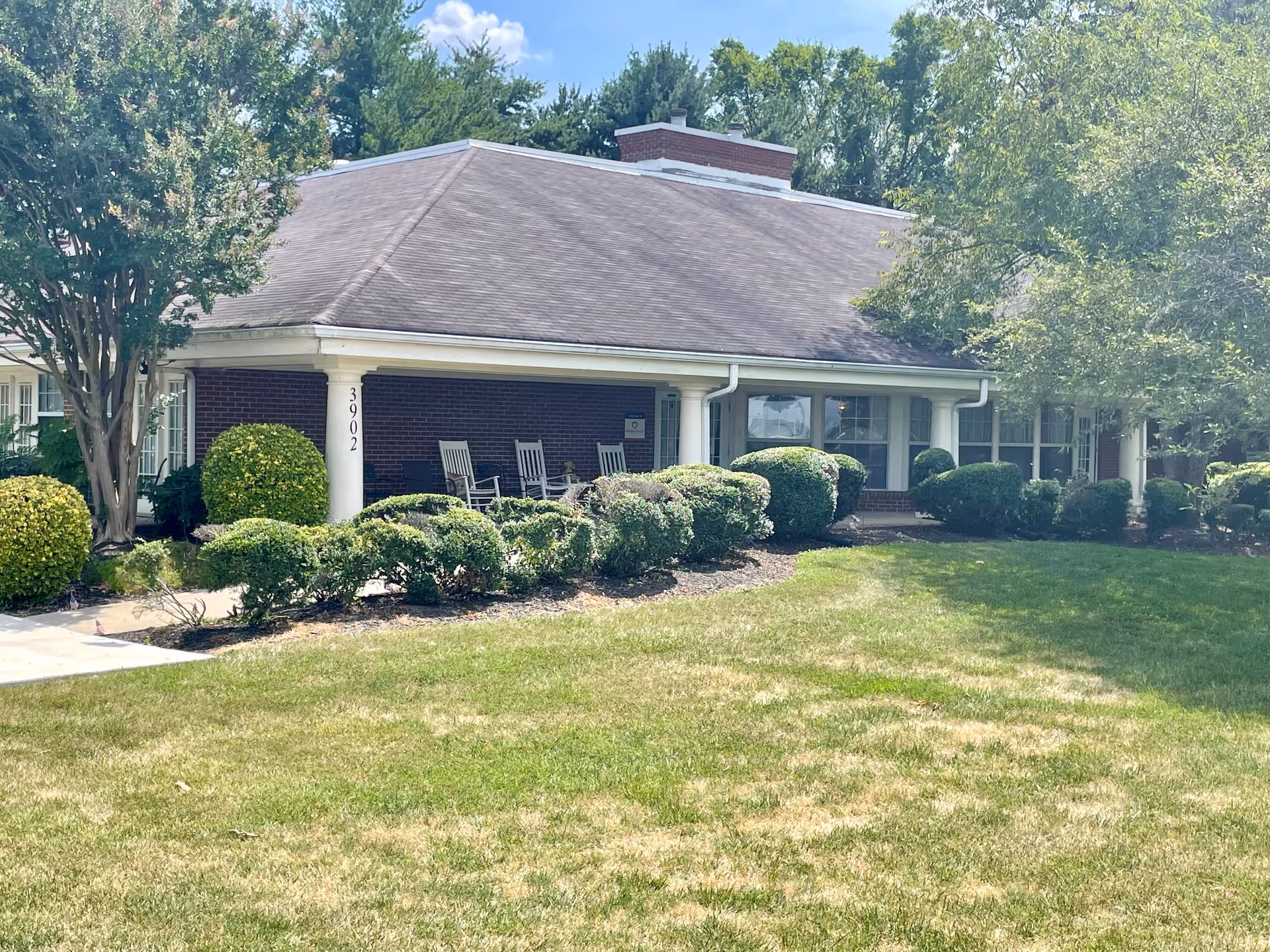 Exterior view of a single-story brick building with a covered porch featuring rocking chairs. The building is surrounded by neatly trimmed bushes, trees, and a well-maintained lawn under a clear blue sky.