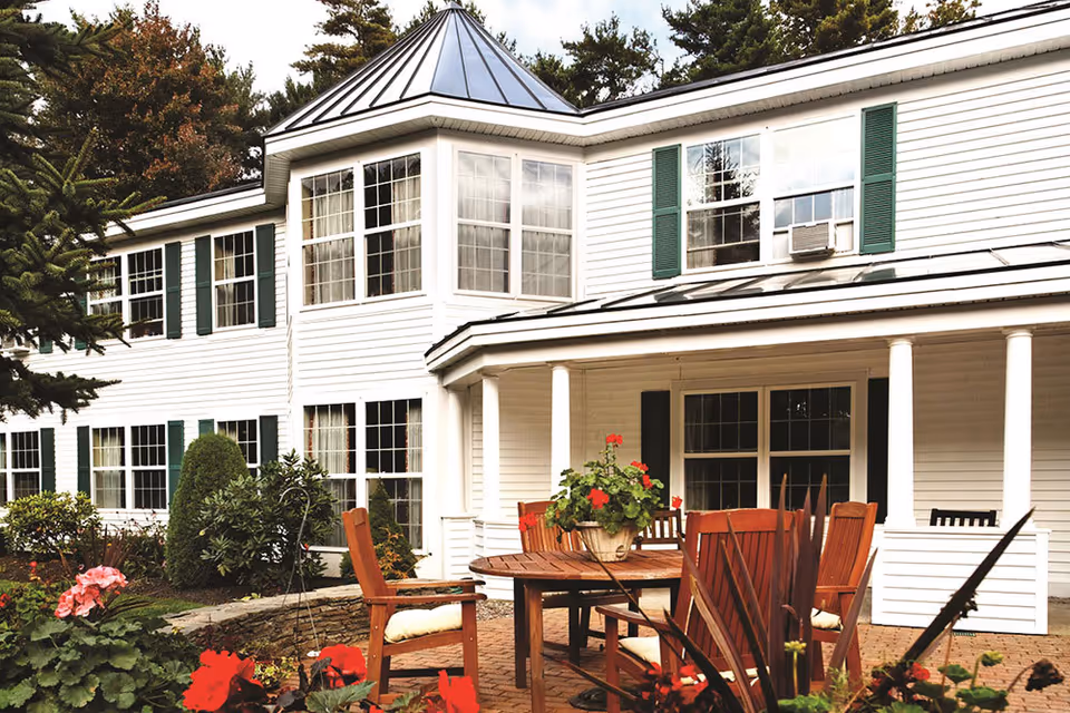 Front exterior of a white two-story building with a covered porch, patio table and wooden chairs surrounded by potted flowers and landscaping.