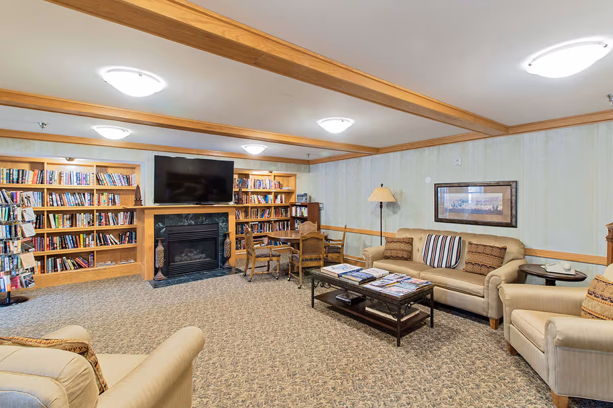 A cozy living room area in an assisted living facility featuring beige sofas and armchairs with patterned cushions, a coffee table with magazines, a floor lamp, and a wall-mounted TV above a fireplace. Built-in wooden bookshelves filled with books line the back wall, and a small table with chairs is placed near the fireplace.