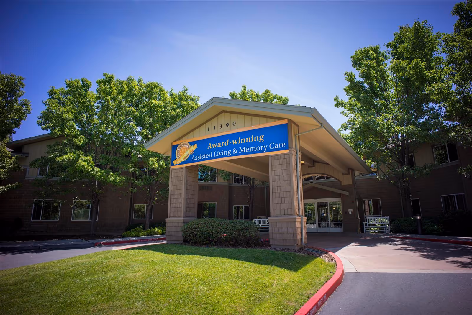 Front entrance of a senior living facility with trees and a blue banner sign.