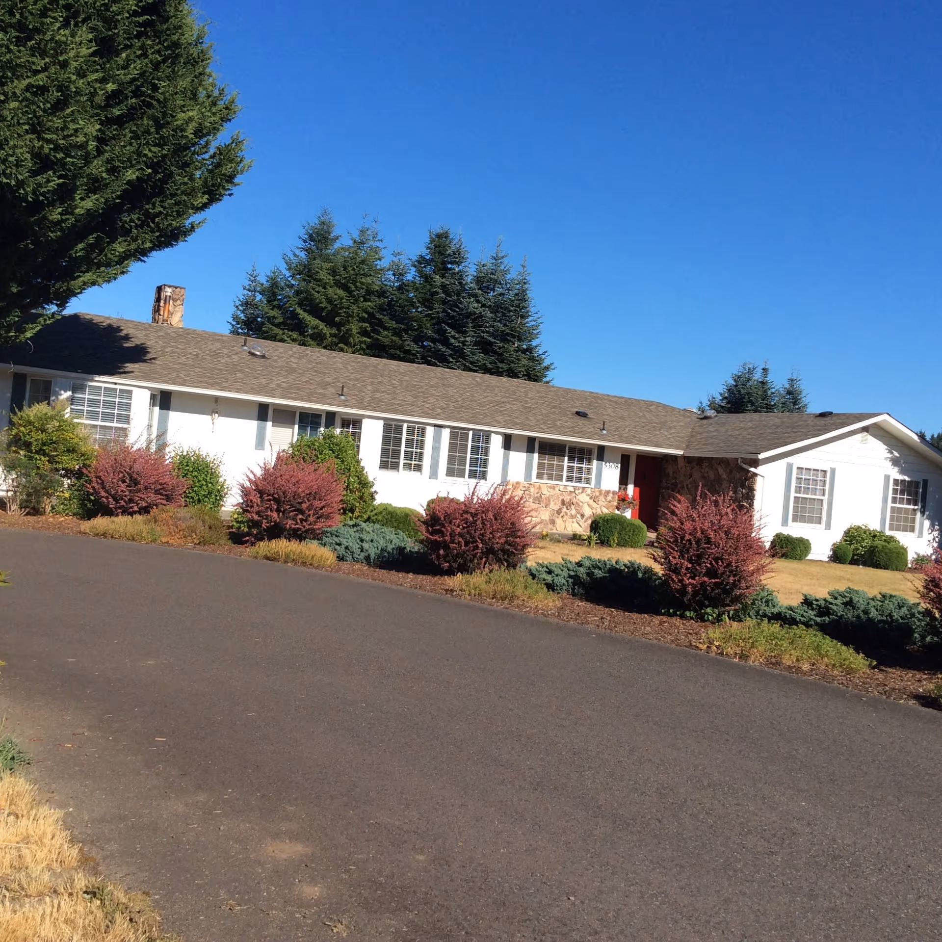 Single-story white building with stone accents, red entrance door, and manicured shrubs along the driveway.