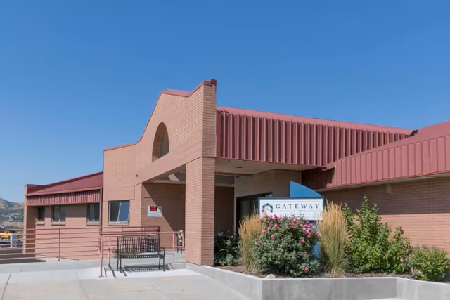 Exterior view of Gateway Transitional Care Center building with brick walls, a red roof, a bench near the entrance, and landscaping with bushes and flowers under a clear blue sky.